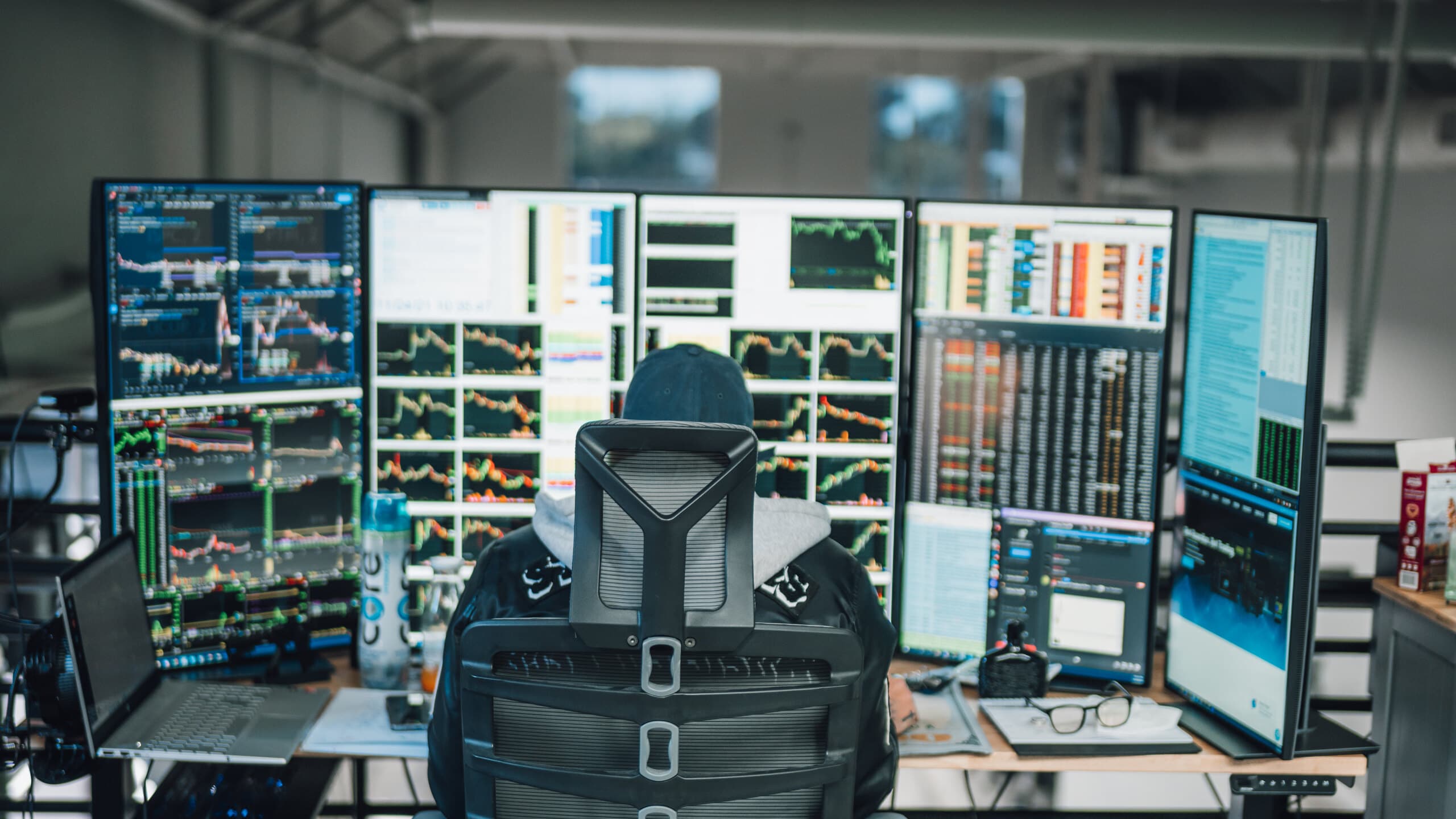 A trader seen from behind at a five-screen rig of charts